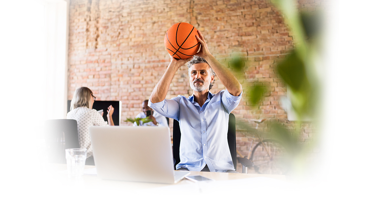 A man in an office with colleagues plays with a basketball at his desk