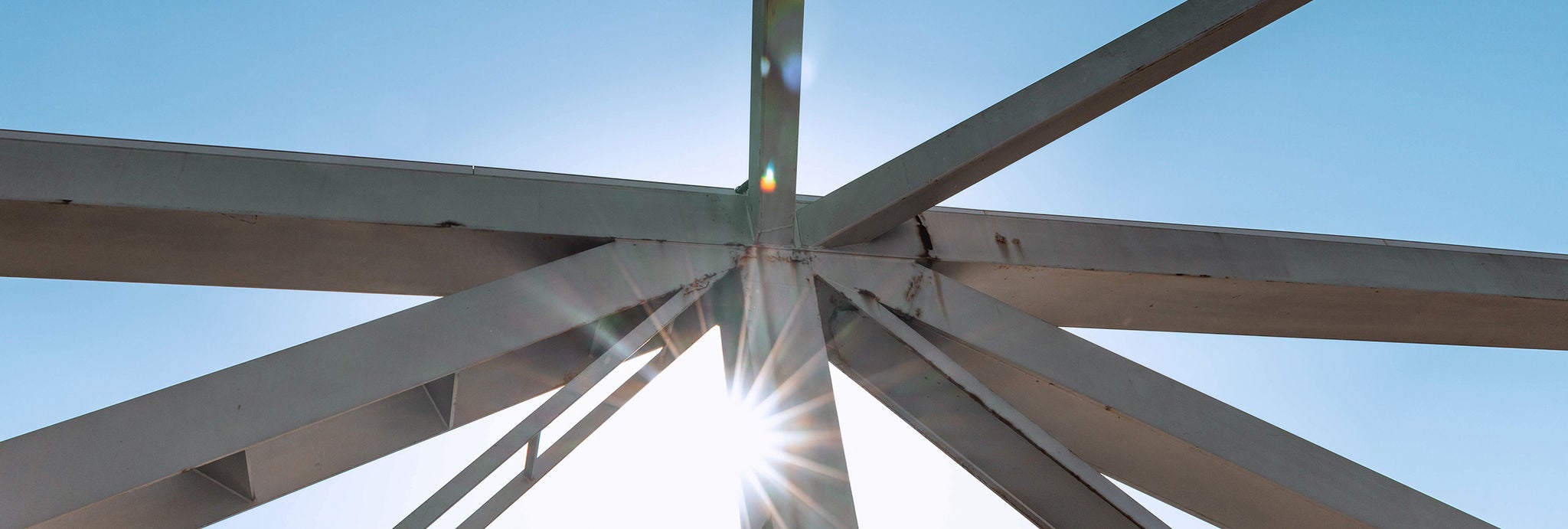 Sunlight shines through intersecting steel beams against a clear blue sky.