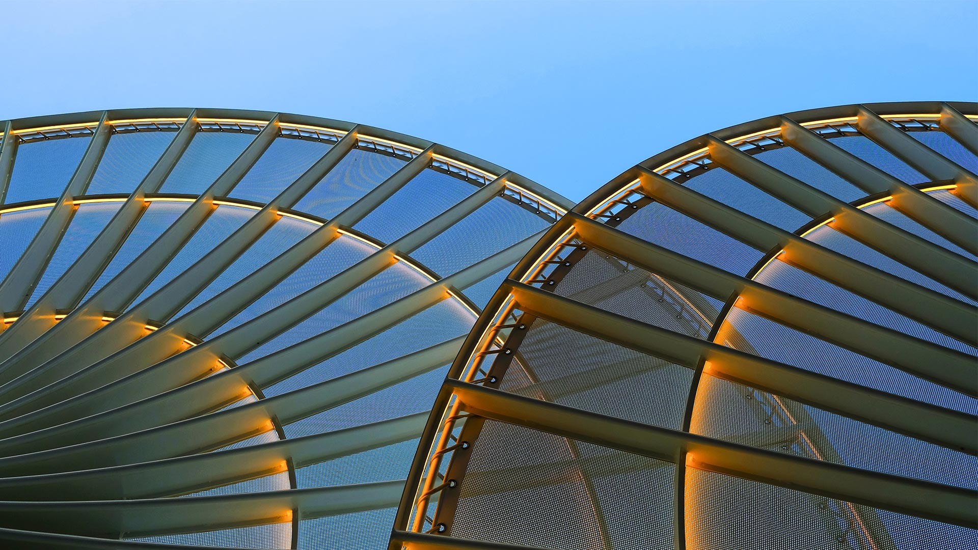 Abstract view of a curved architectural canopy with illuminated metal beams against a blue sky