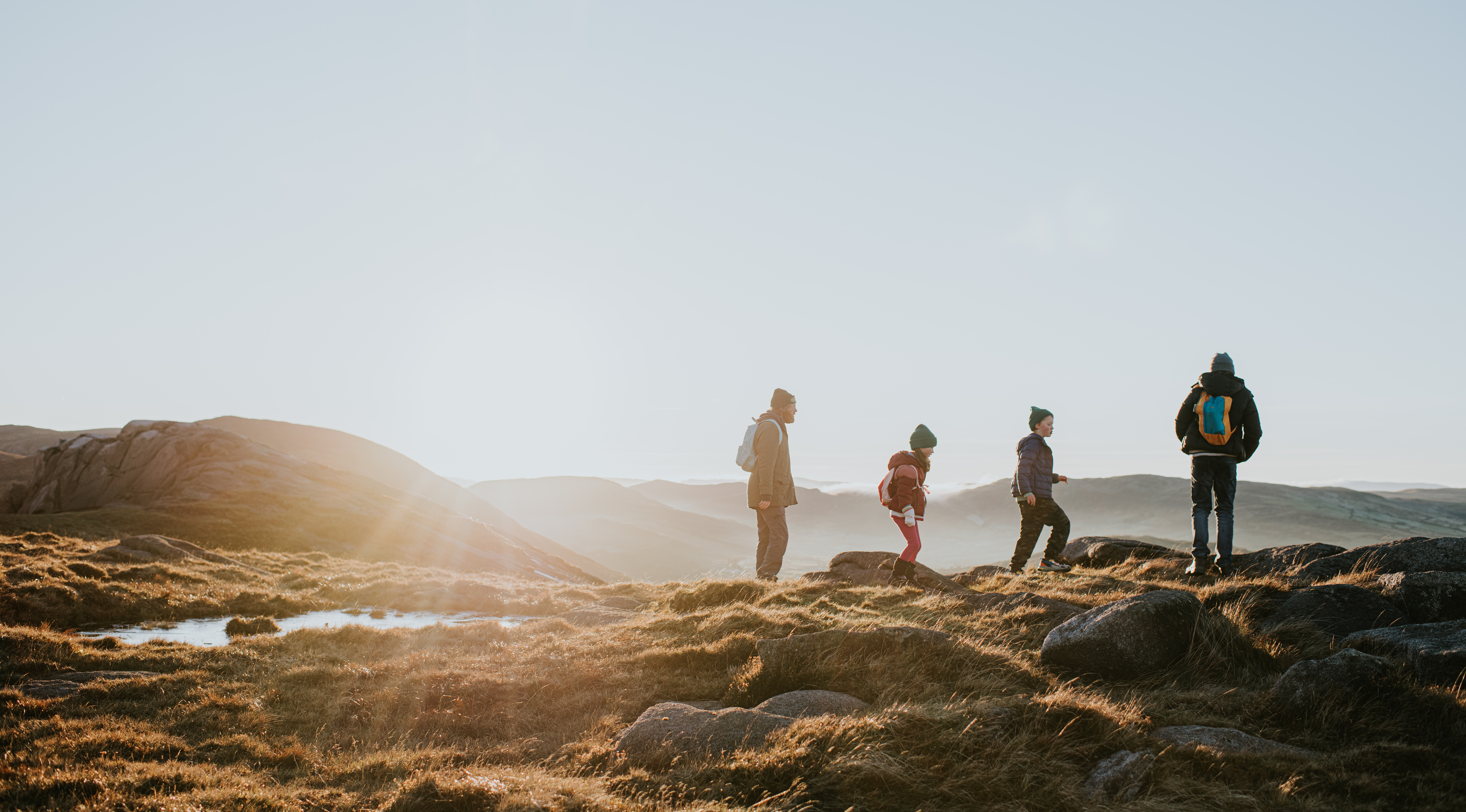 Four people hike along a rocky, grassy hillside at sunrise with mountains