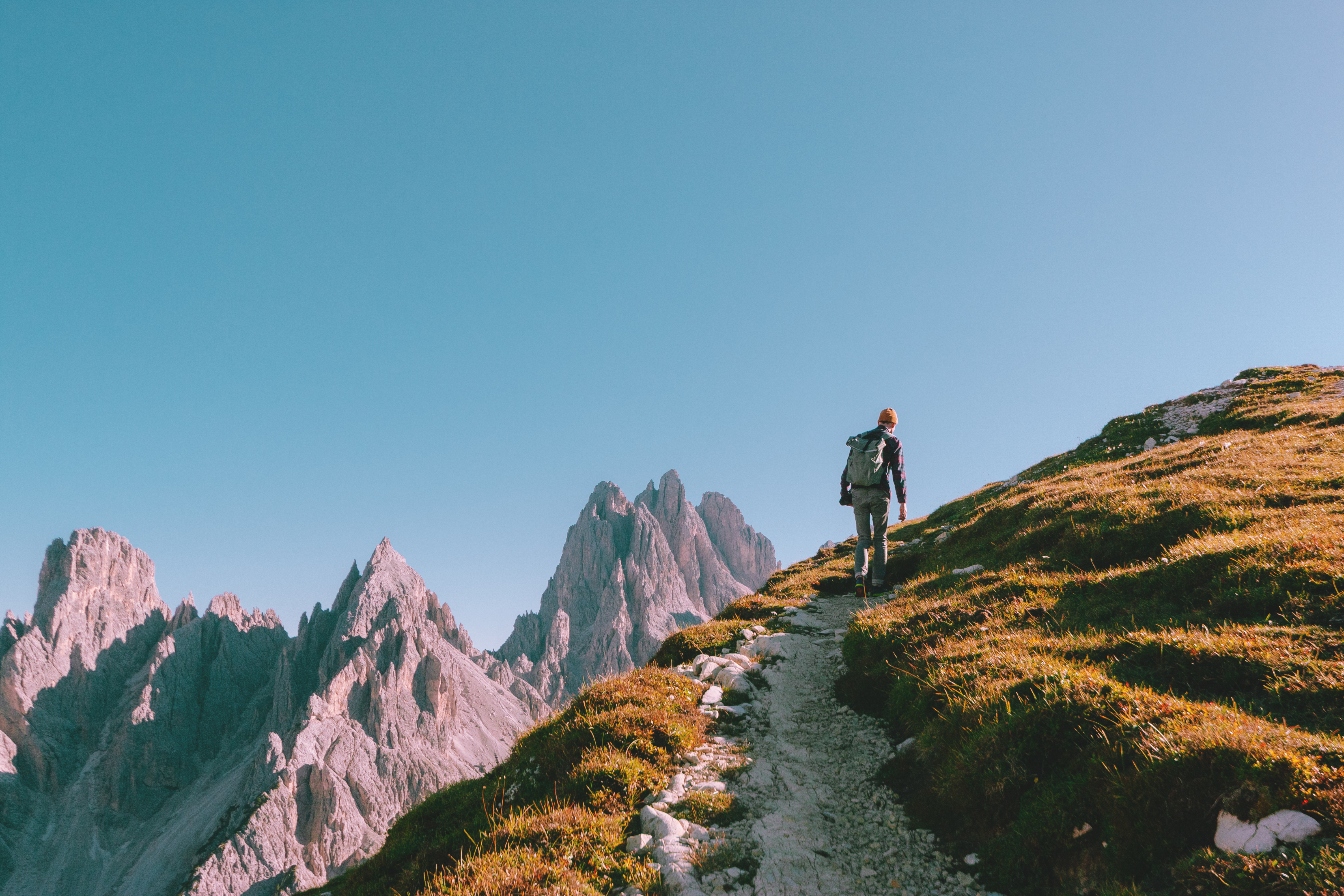A hiker with a backpack walks up a mountain trail on a grassy slope, with rugged alpine peaks in the background under a clear blue sky