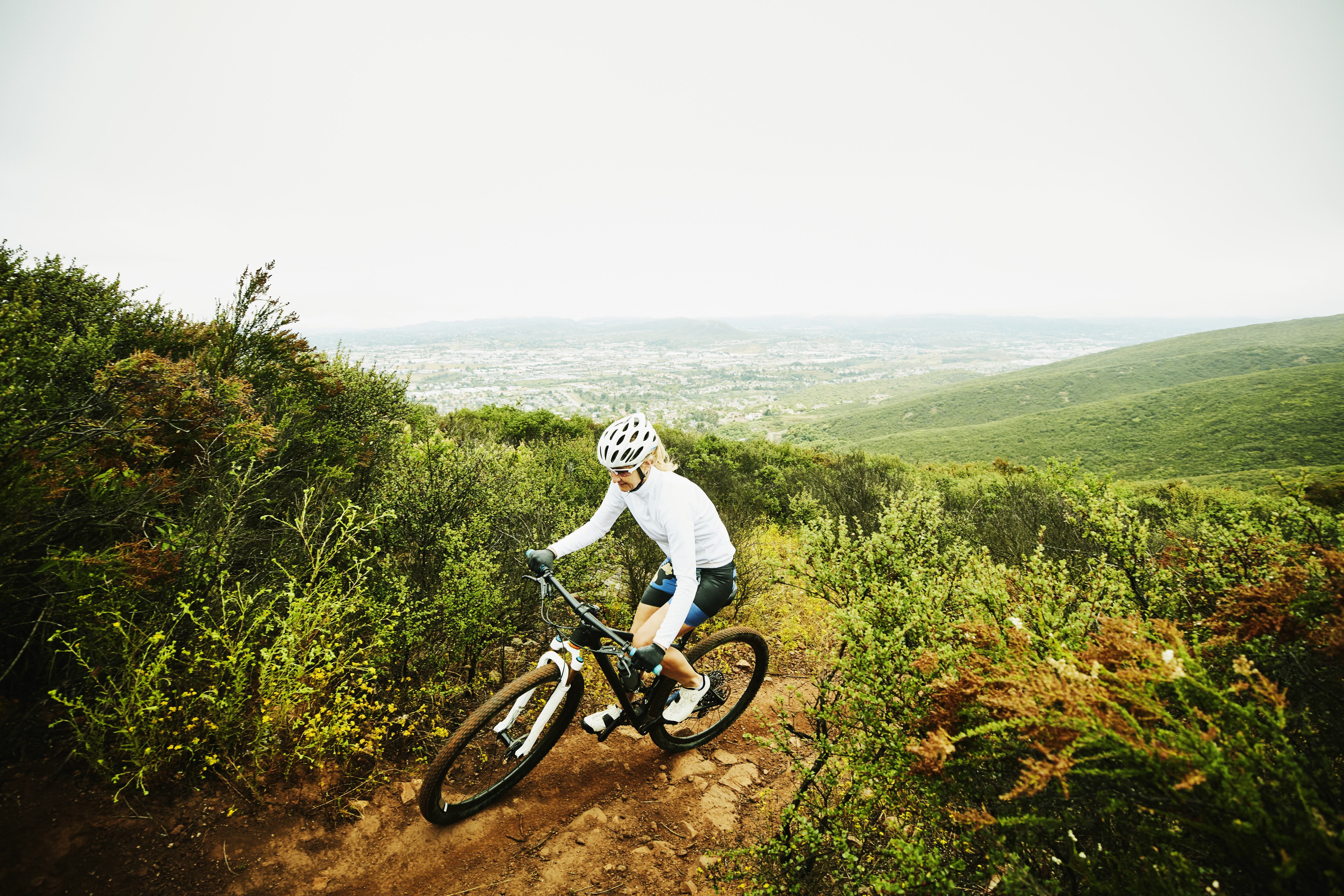 A hiker with a backpack walks up a mountain trail on a grassy slope, with rugged alpine peaks in the background under a clear blue sky