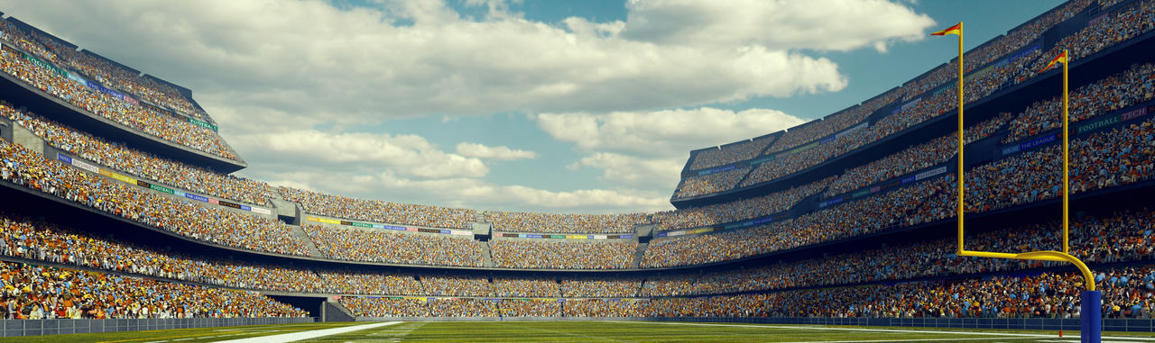 A wide angle panoramic image of a outdoor american football stadium full of spectators under blue sky. The image has depth of field with the focus on the foreground part of the pitch. The view from back line of the field.
